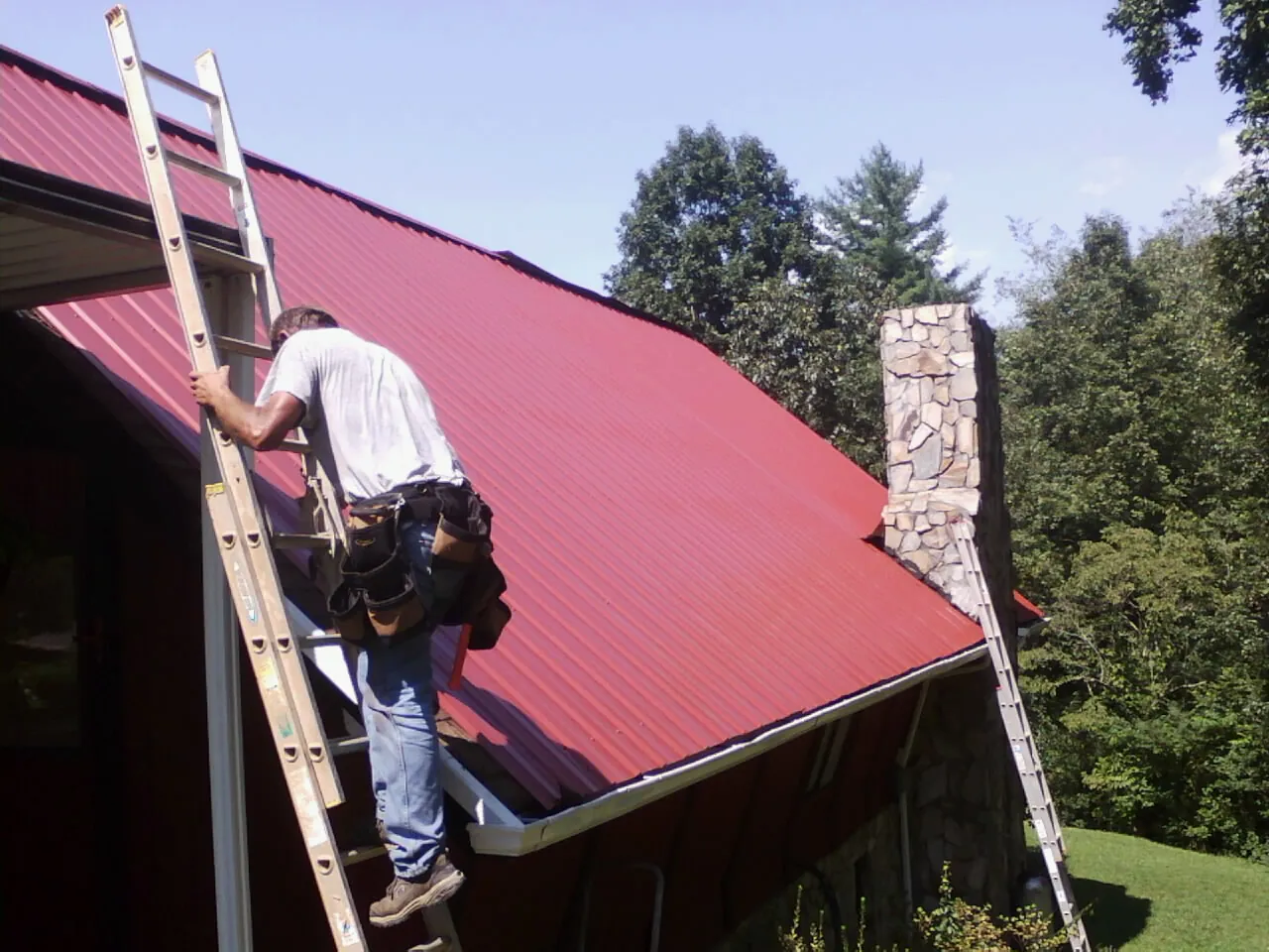 Roofer on a ladder beside a red metal roof and stone chimney — Soffit & Fascia Repair by Roofing Brothers in North Granby